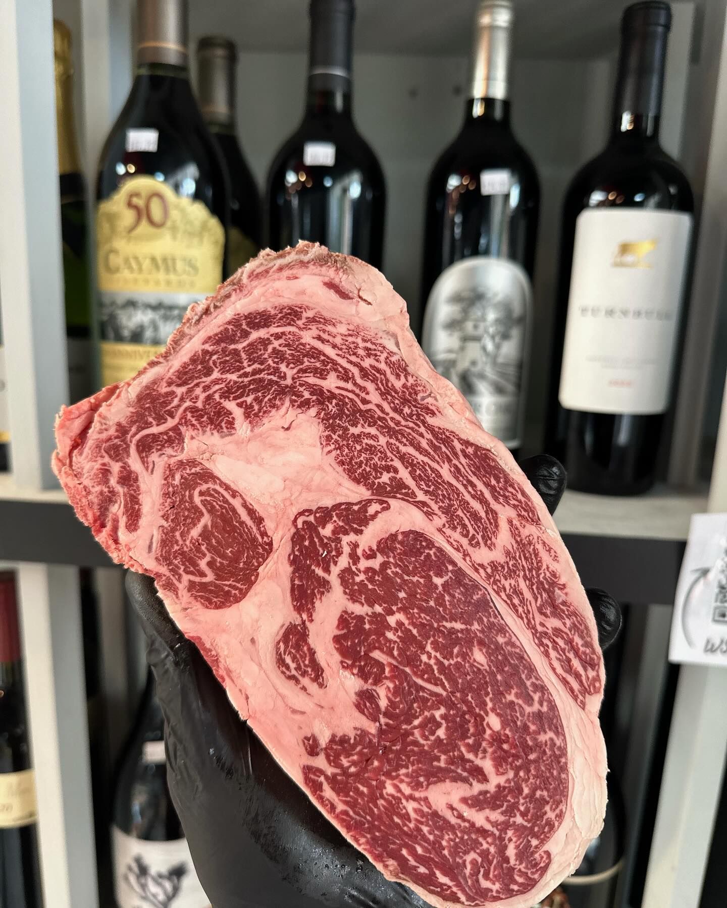 Hand holding a raw, marbled ribeye steak in front of a shelf of wine bottles.