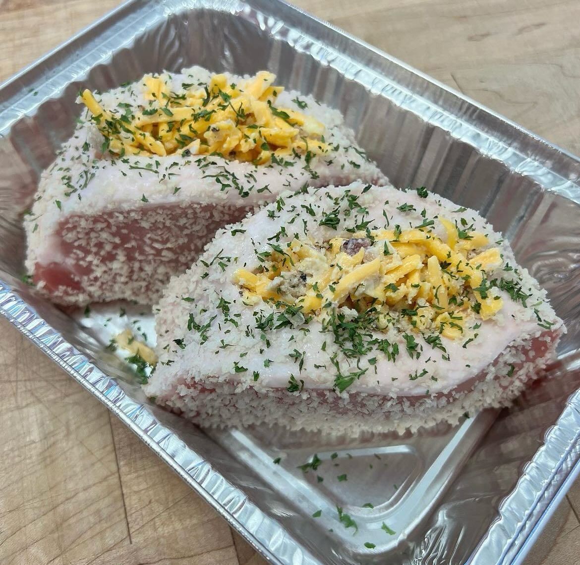 Two breaded pork chops topped with shredded cheese and parsley in a foil tray.