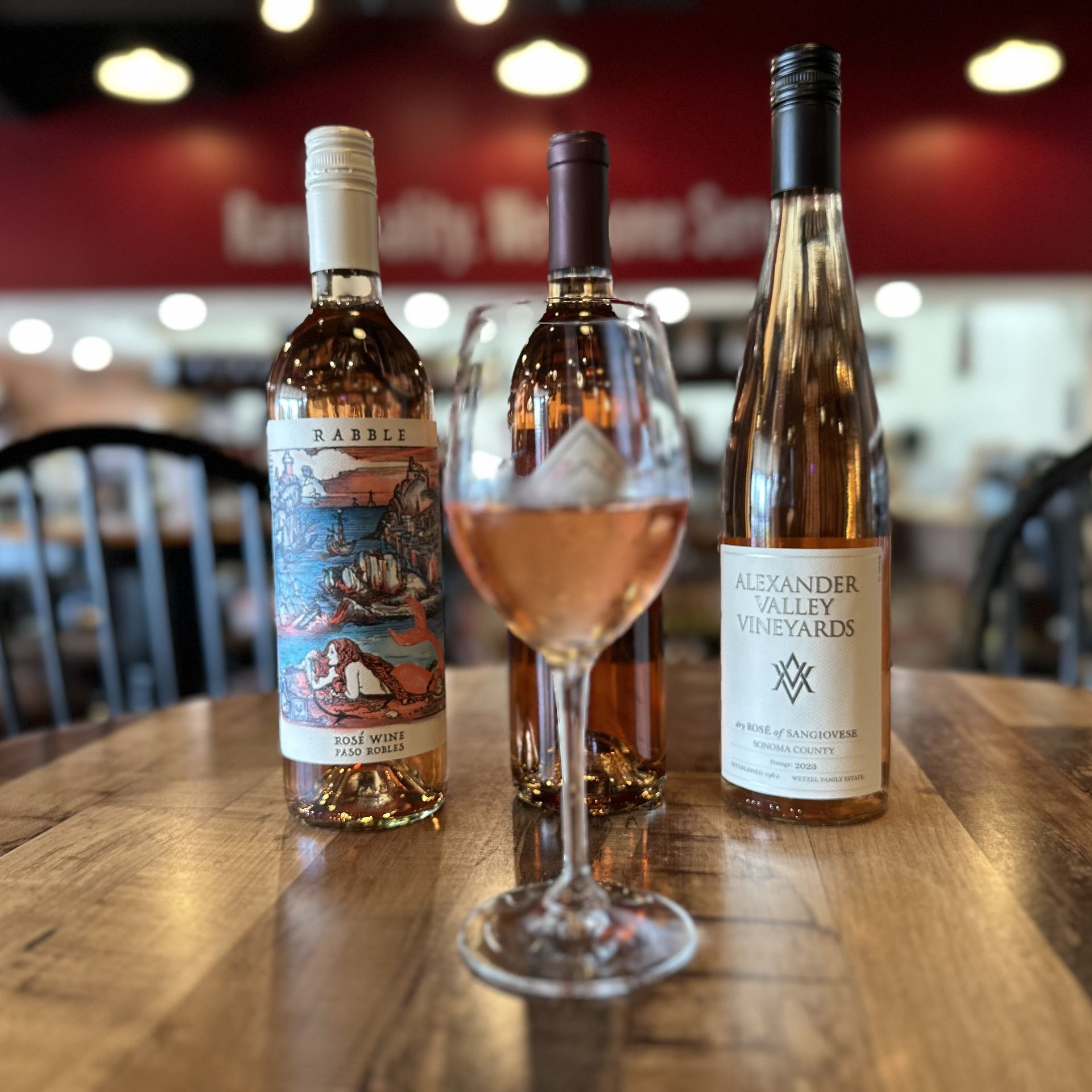 Three bottles of rosé wine and a filled glass on a table; shop interior.