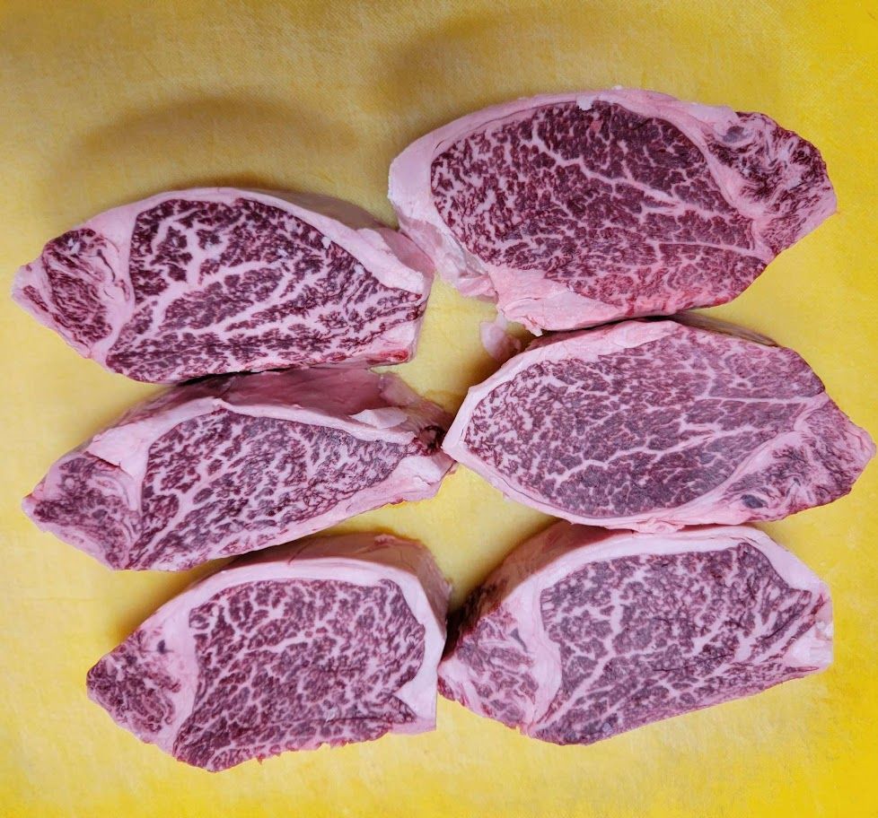 Six raw, marbled steaks on a yellow surface, showing intricate fat patterns and red meat.