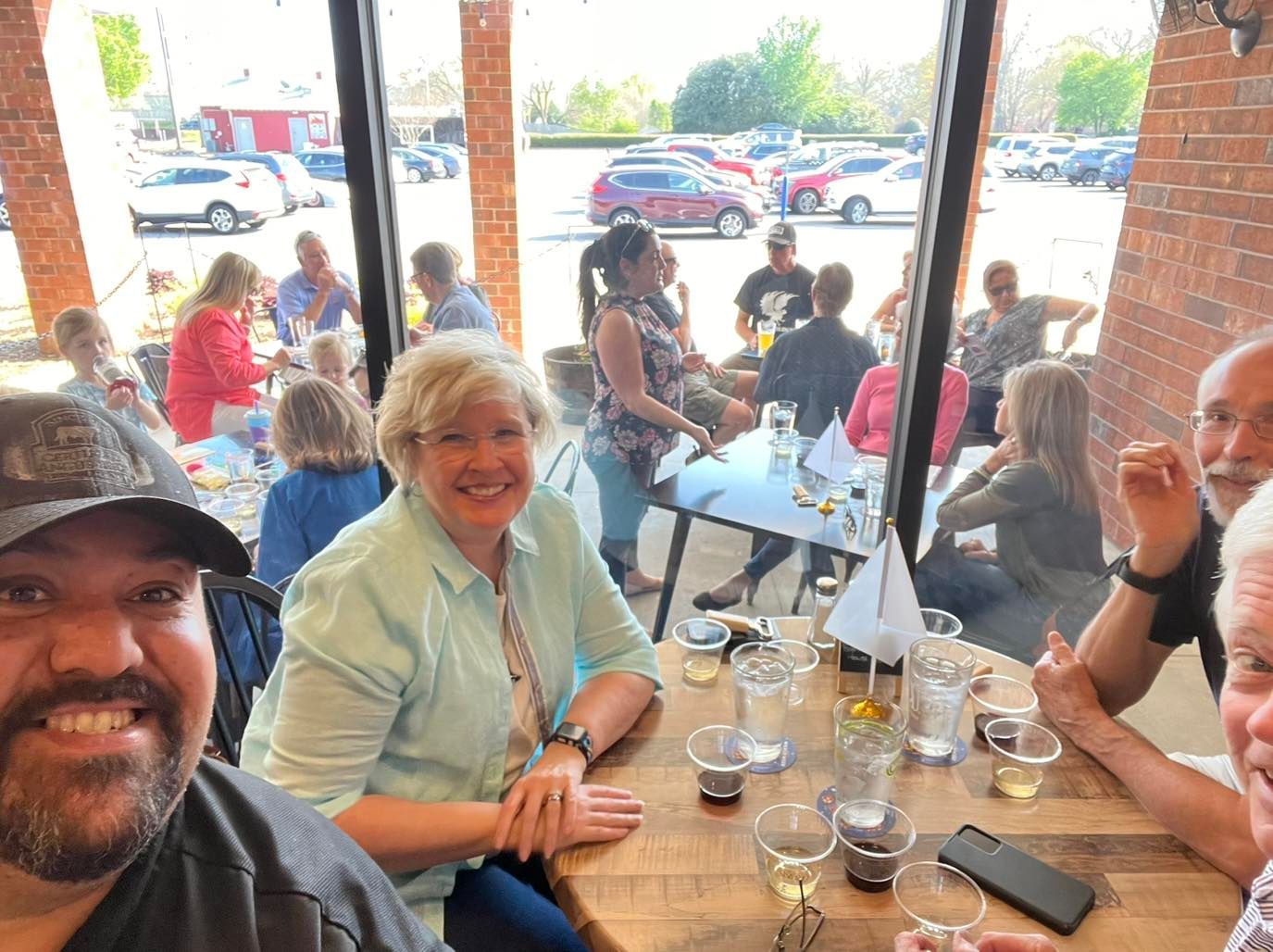 People at outdoor brewery table; smiling woman, beers, window view of parking lot.