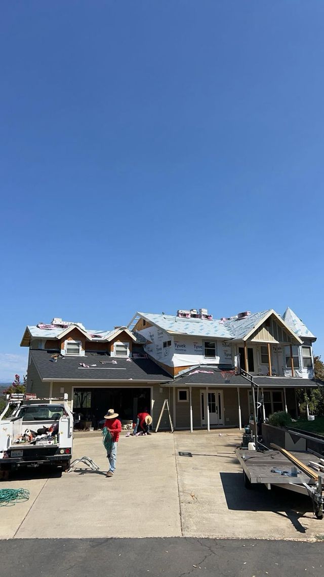 Damaged house with roof damage and debris, workers and equipment in the driveway under a clear blue sky
