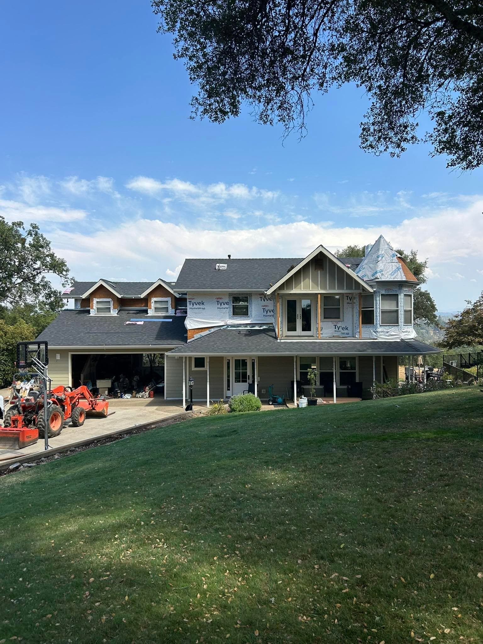 Large gray house with porch on a grassy hill under a blue sky, with red lawn equipment at left.