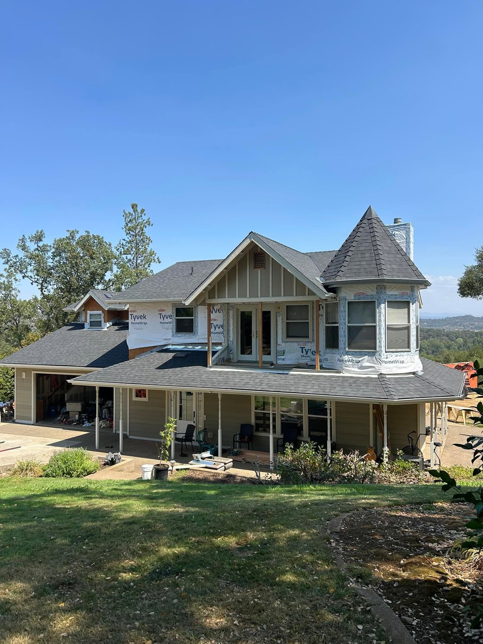 Large white house under construction with porch and gabled roof, set on a grassy lot under a blue sky