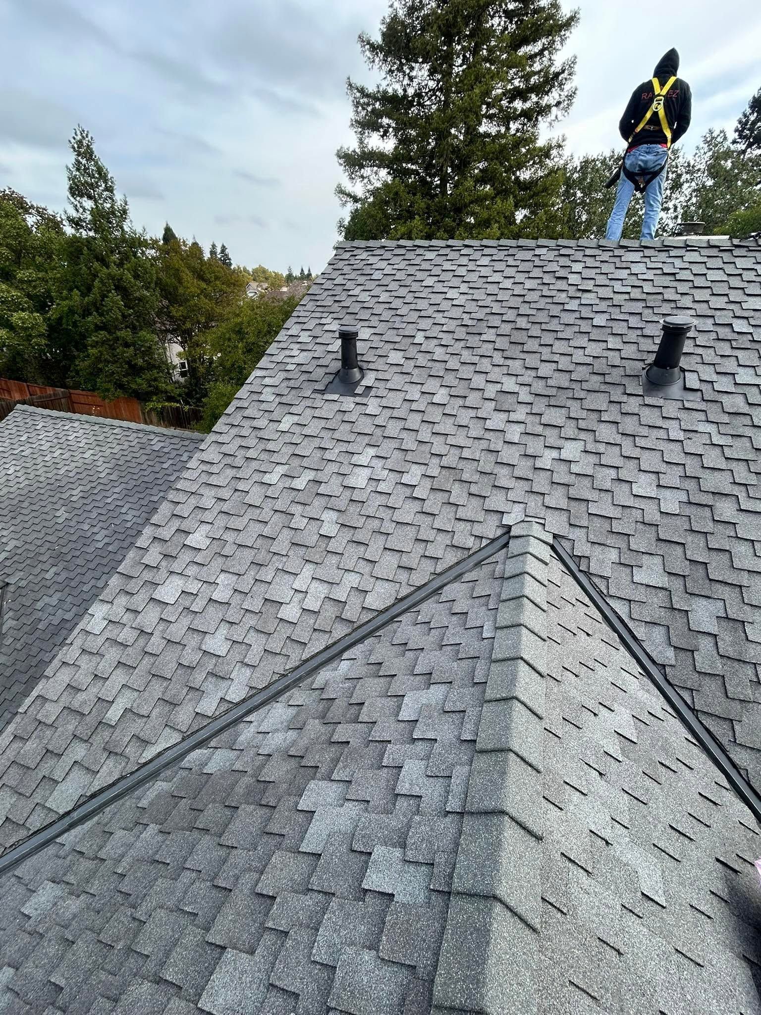 Worker standing on a gray shingled roof near a chimney, with trees in the background