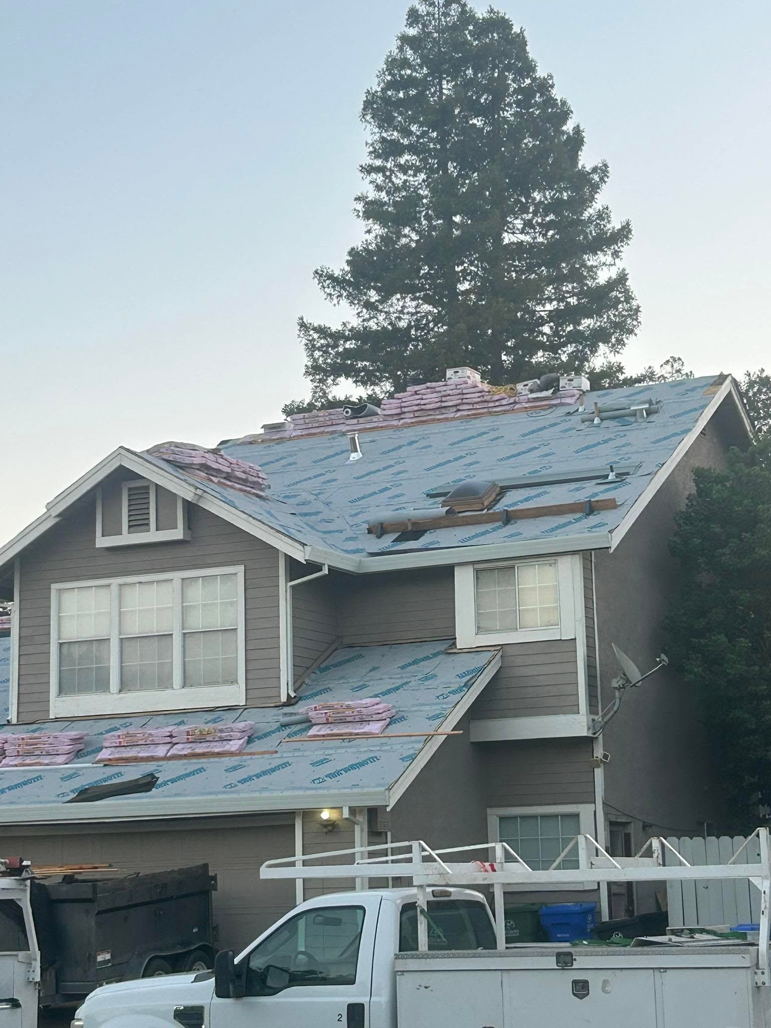 Two-story house with severe roof damage, covered in torn blue tarp, with a large tree behind it