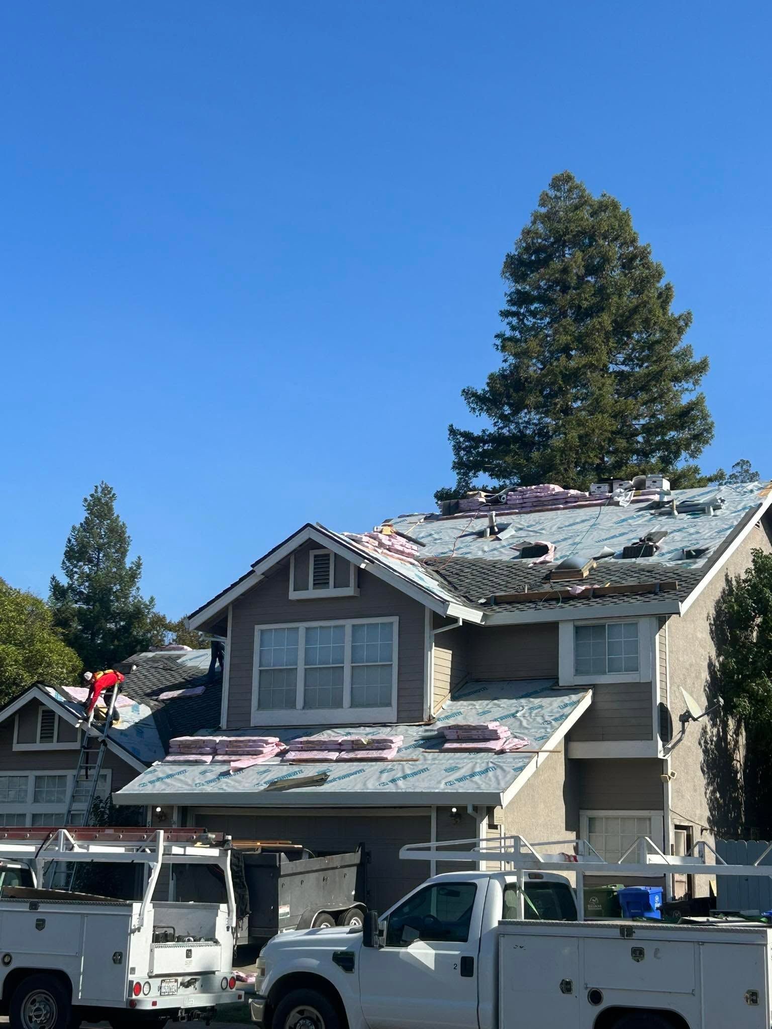 Damaged house roof with debris and patches, with work trucks parked outside under a clear blue sky
