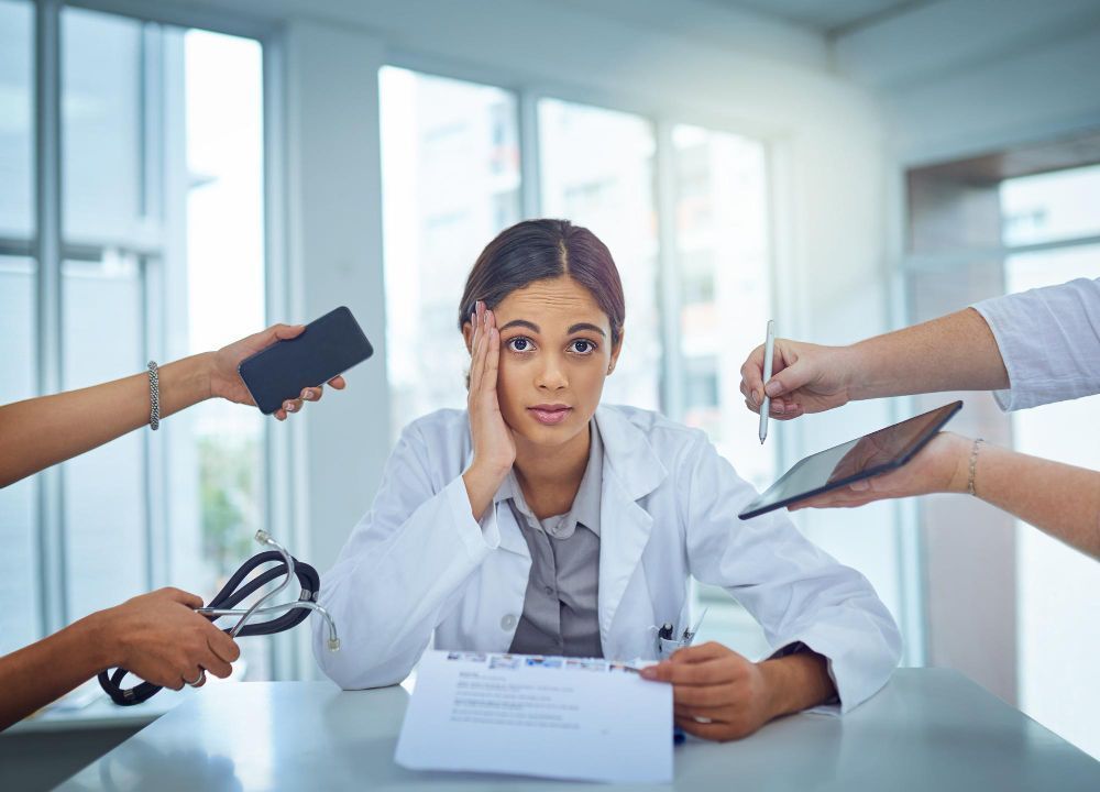 Doctor looks stressed, surrounded by hands holding phone, pen, tablet, and stethoscope at a desk.