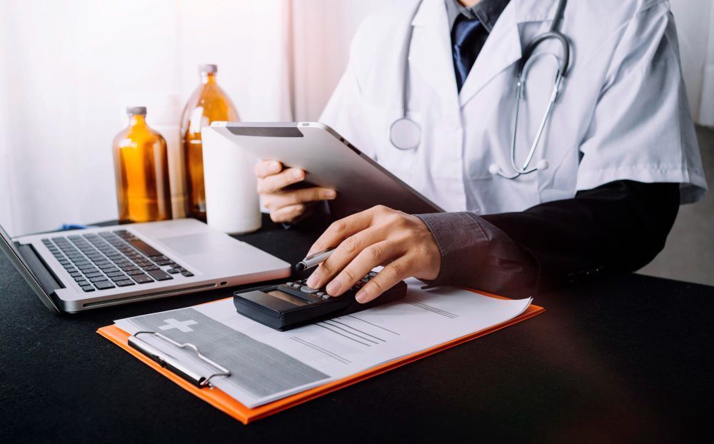 Doctor in lab coat using a calculator and tablet; medical documents on desk.