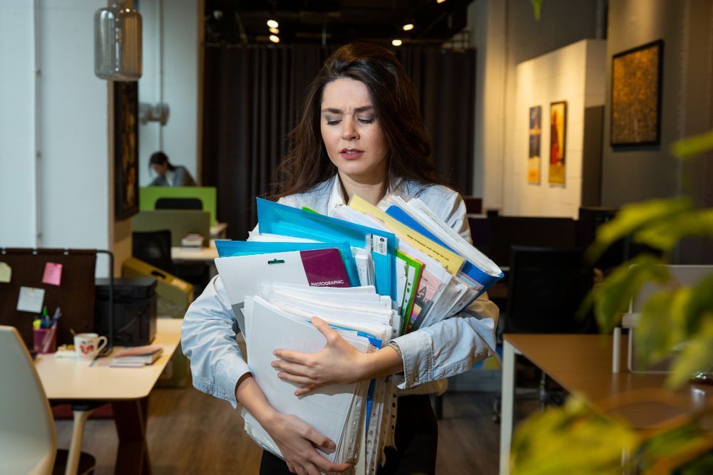 Woman in office carrying a stack of papers, looking stressed.