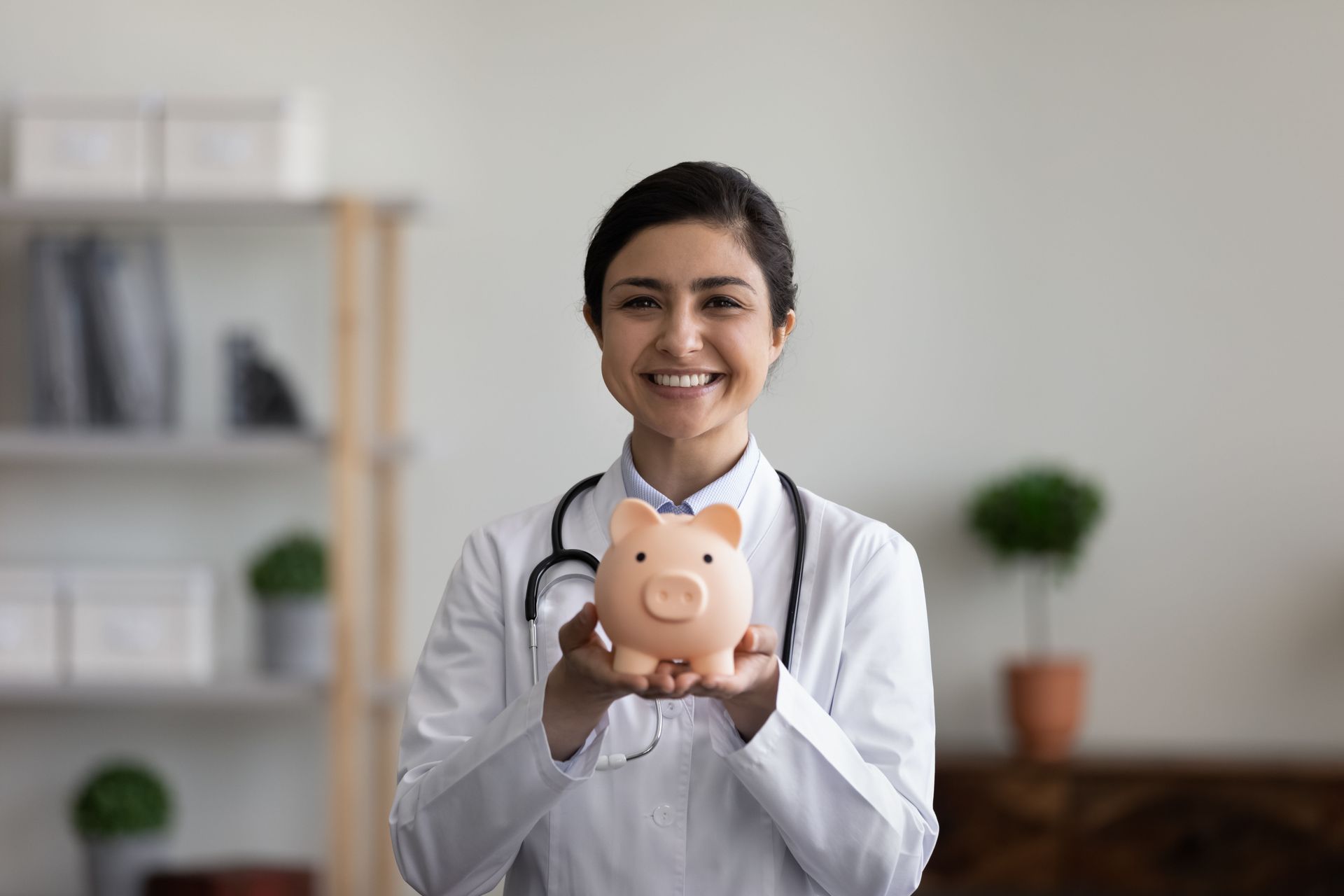 Smiling doctor holding a pink piggy bank, stethoscope, indoor setting.
