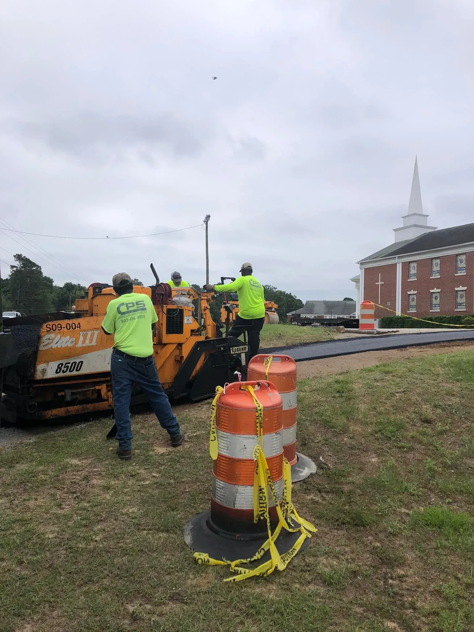 Road crew in neon vests working beside orange construction equipment on a roadside under a cloudy sky