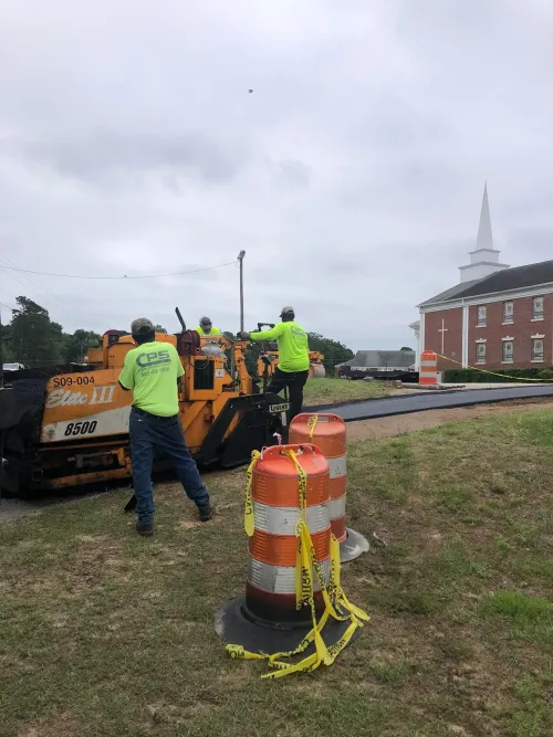 Workers repair a road near orange barrels and heavy machinery on a cloudy day.
