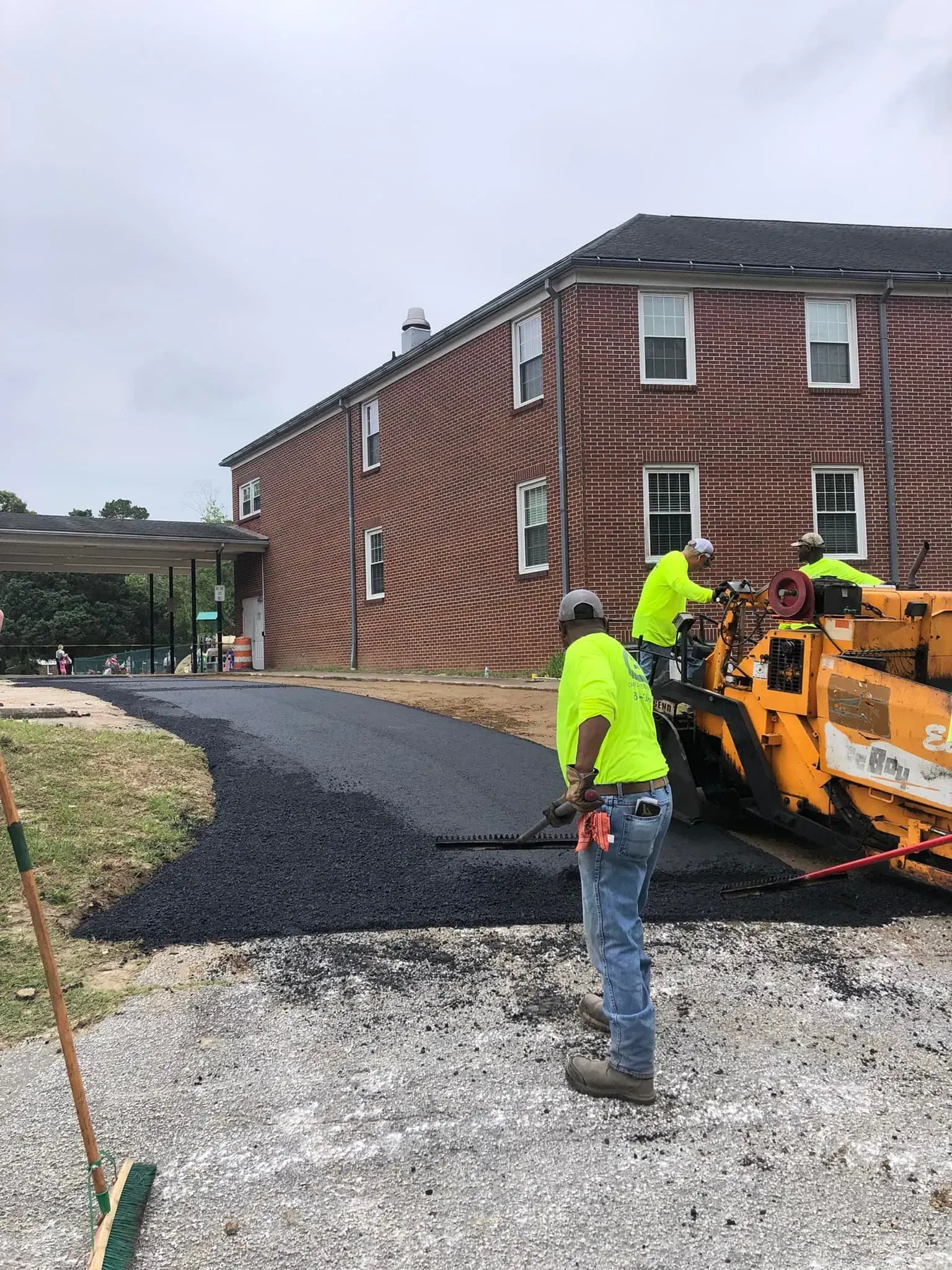 Construction workers paving a gravel path beside a brick building with an asphalt machine.