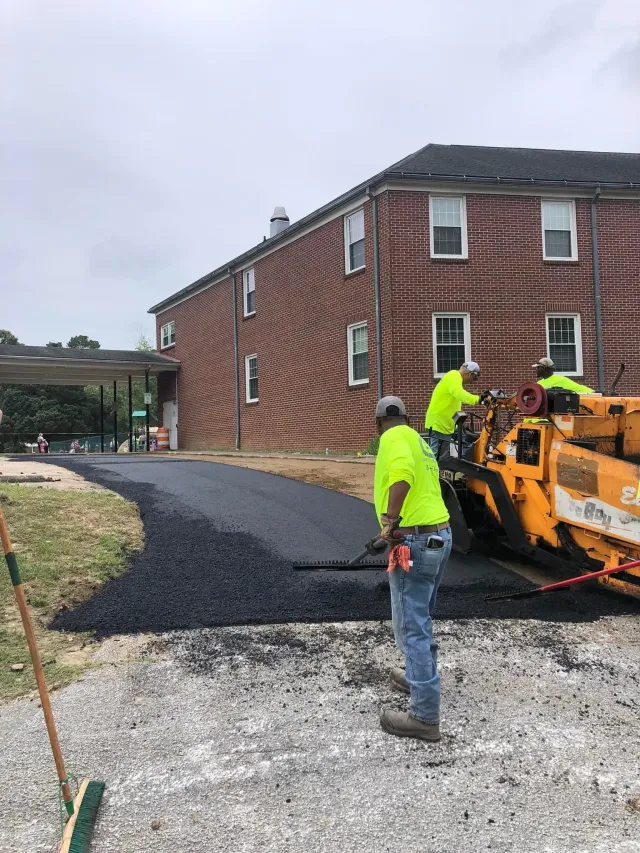 Workers using a road machine to spread gravel beside a brick building under construction