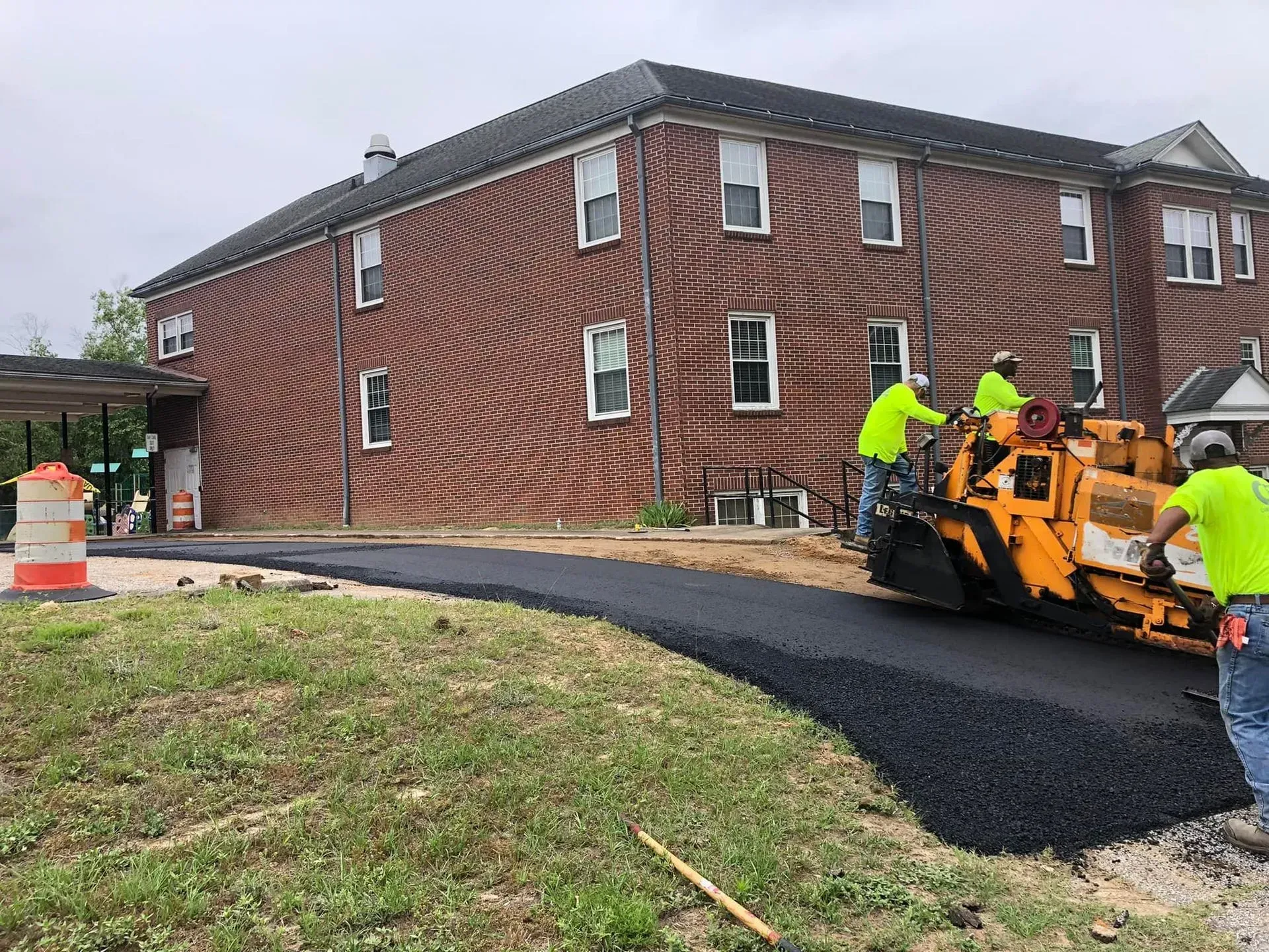 Workers paving a black driveway beside a red brick building on an overcast day