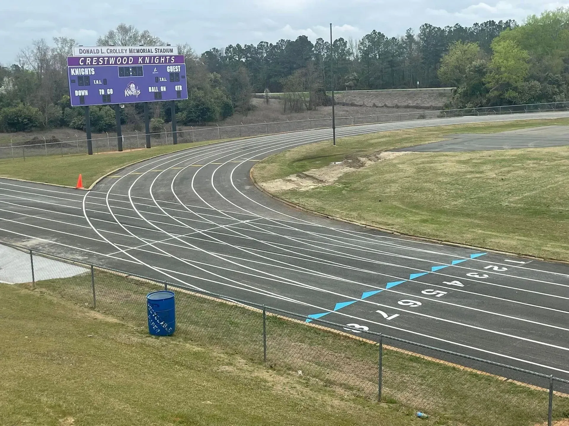 Empty outdoor running track with lane markings, a purple scoreboard, and trees in the background.