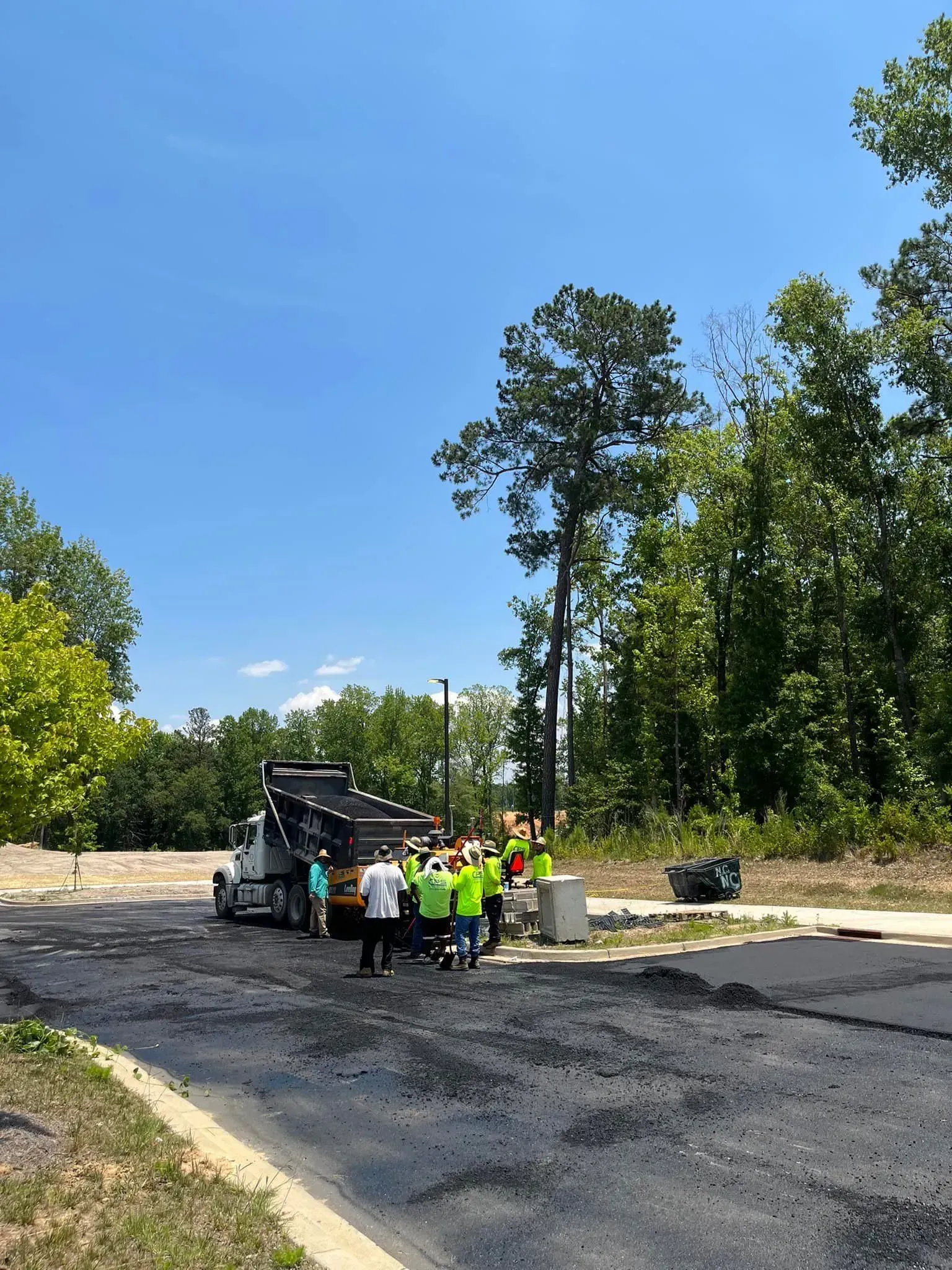 Workers in neon vests stand beside paving equipment on a newly paved road under a blue sky.
