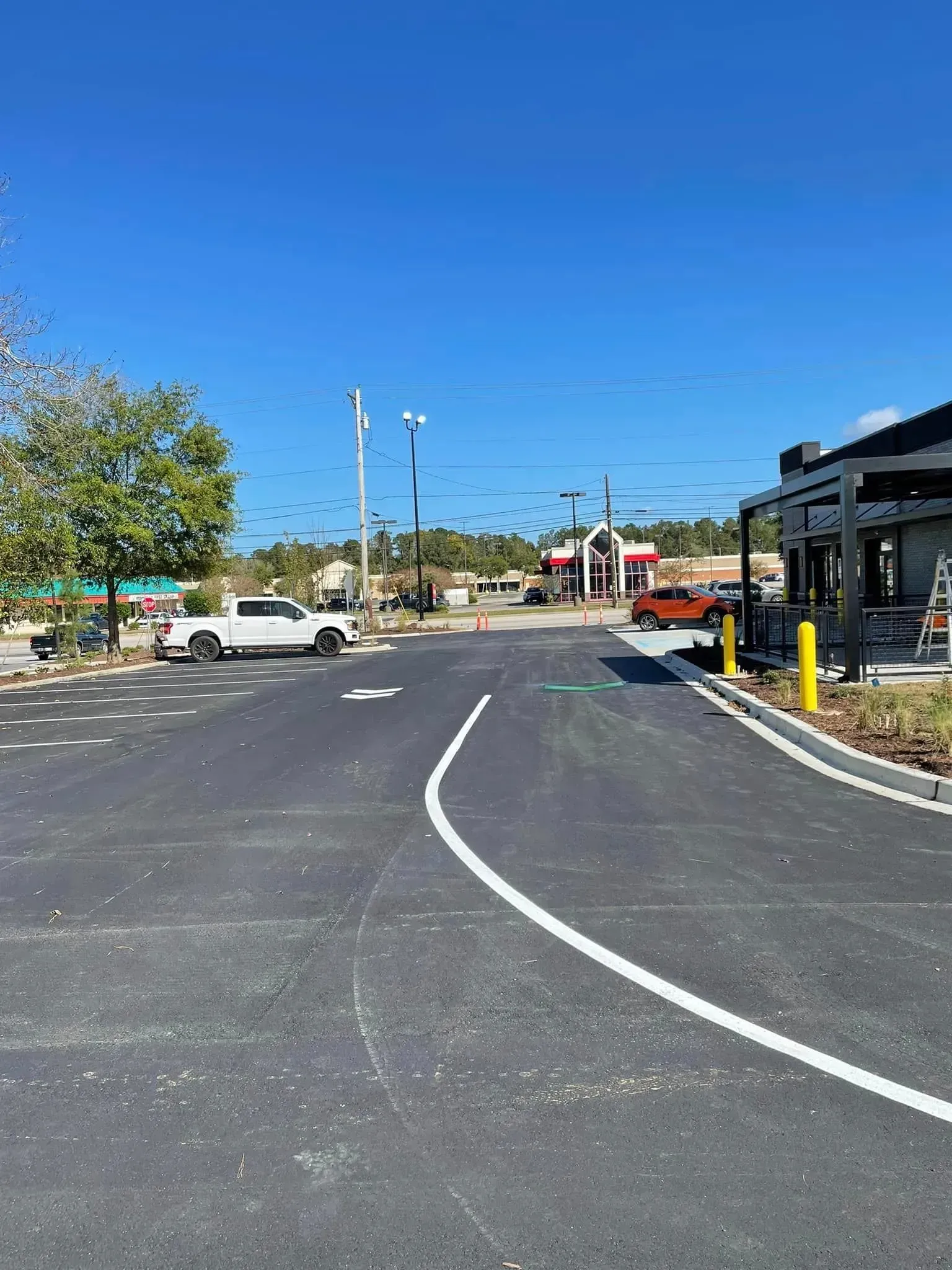 Curved road in a sunny parking area with parked cars, streetlights, and a building on the right.