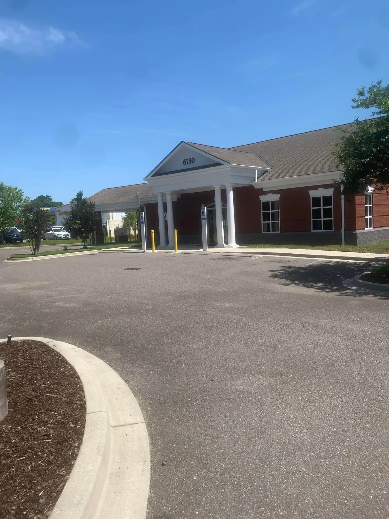 Brick building entrance with white columns, covered drop-off area, and curved driveway under a clear blue sky