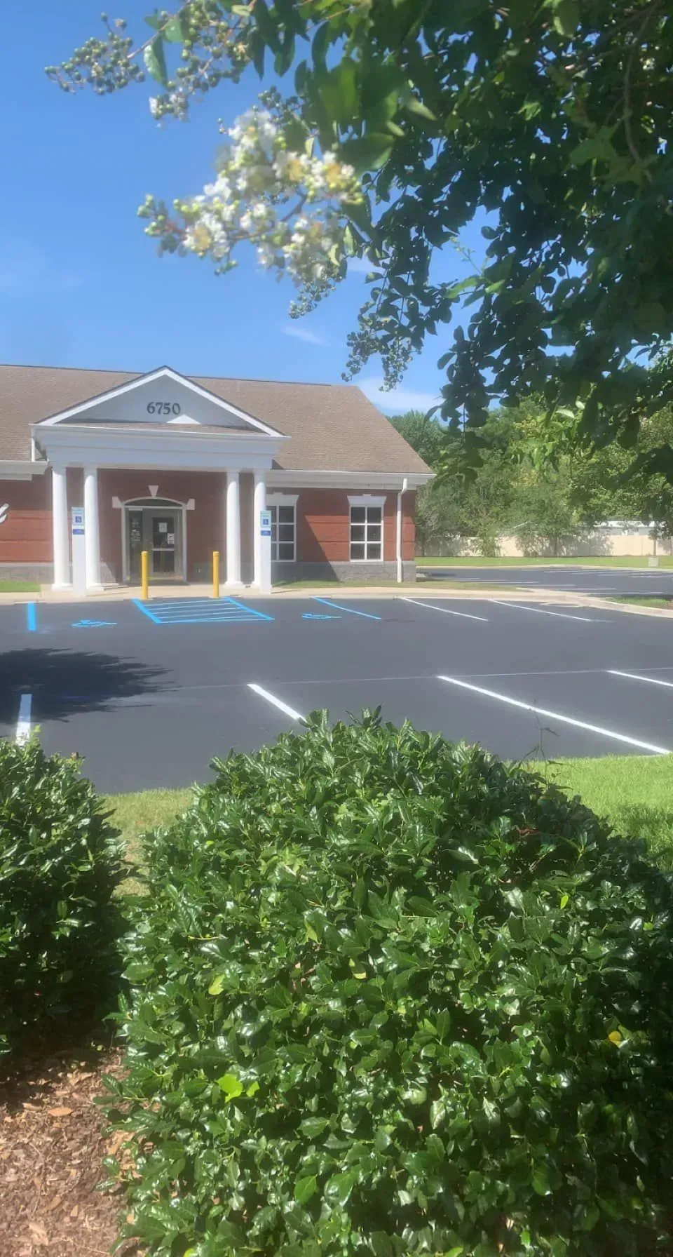 Brick building with white columns beside an empty parking lot, framed by trees and bright blue sky.