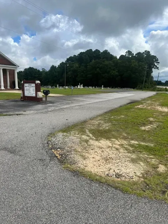 Gravel driveway beside a brick building, leading past a sign toward a grassy field and trees under cloudy sky