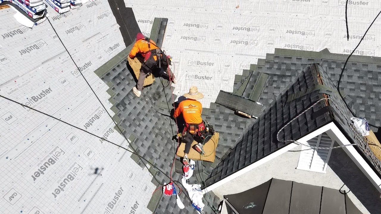Two roofers in orange vests installing shingles on a house roof.
