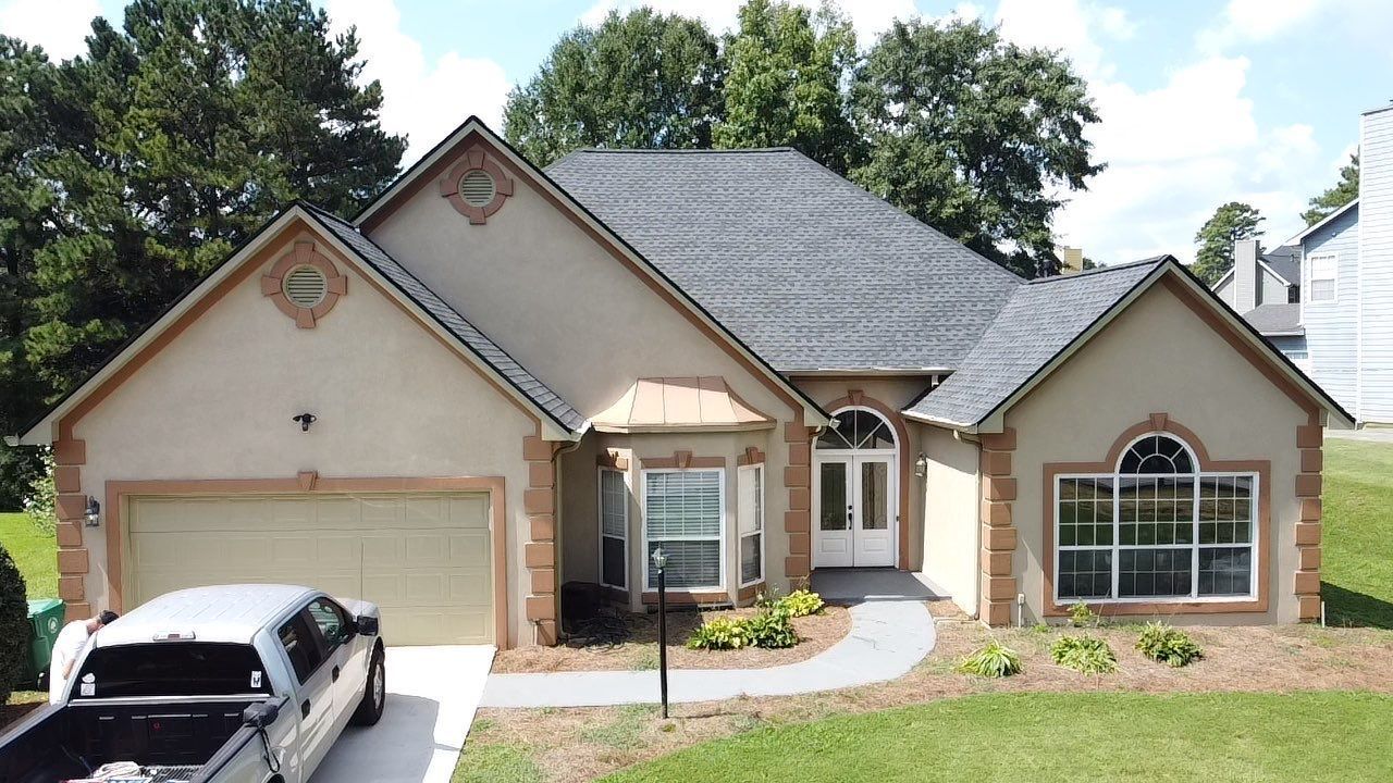 Tan house with black roof, attached garage, and landscaping. A car is parked in the driveway.