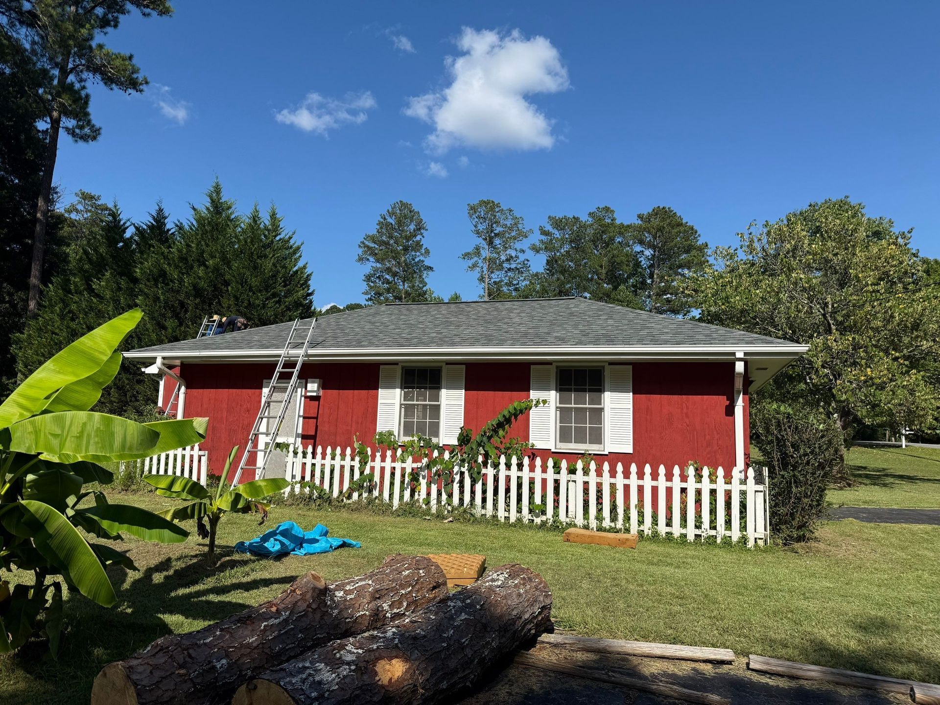 Red house with white picket fence, ladder on roof, logs in the foreground, and a blue sky.