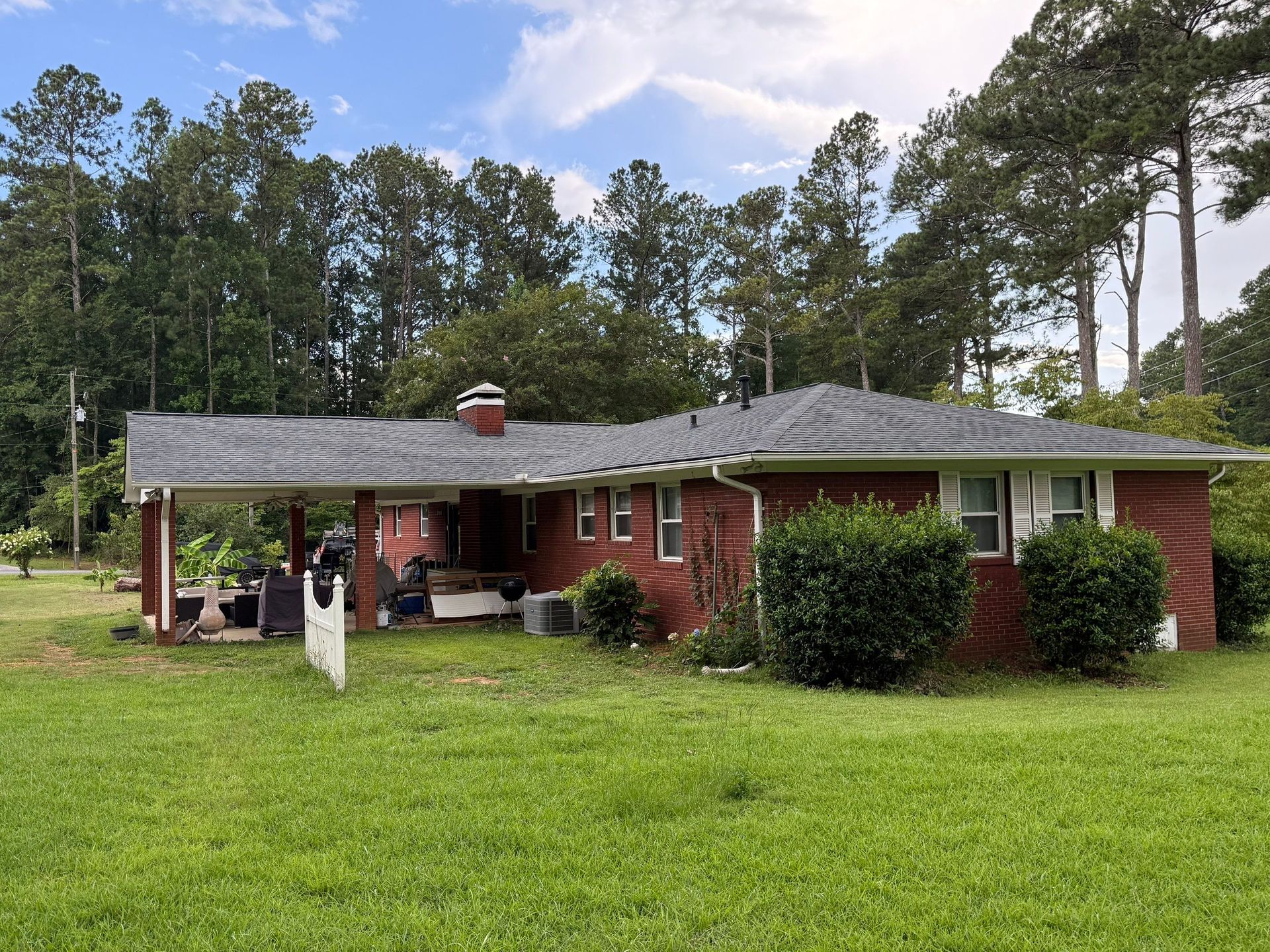 Red brick house with a carport and a green lawn, set against a backdrop of trees and a cloudy sky.