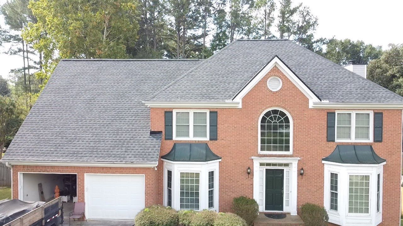 Brick house with a gray roof and white garage door. Trees in the background, blue shutters.