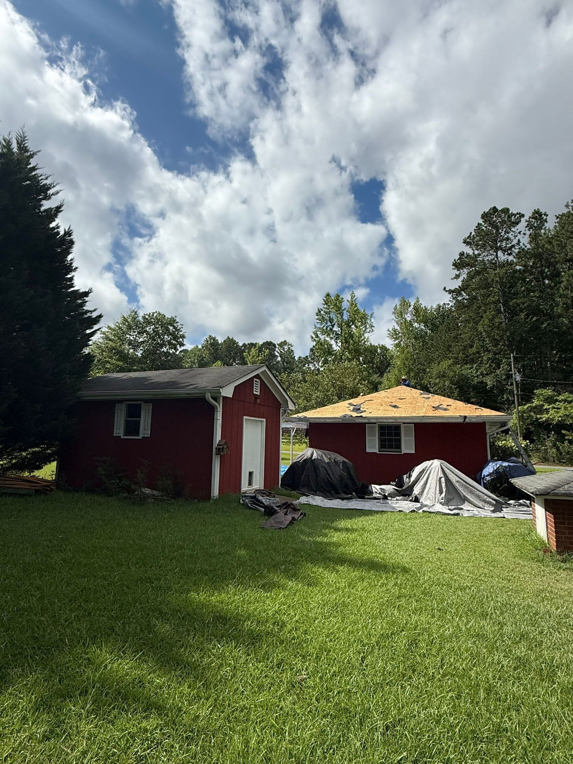 Two red outbuildings under a cloudy sky. One has a new, unfinished roof. Green grass and trees surround them.