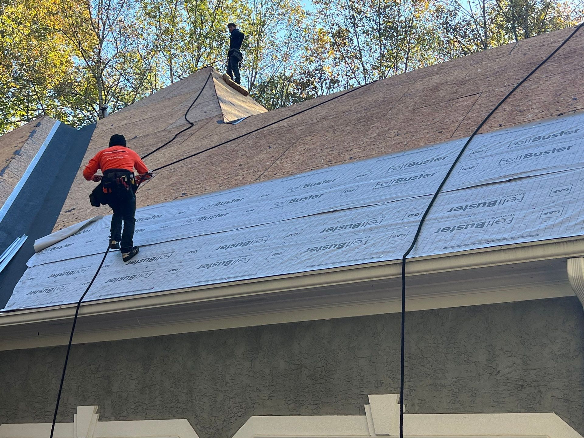 Two roofers working on a house roof; one on the ridge, the other climbing the slope, safety lines visible.