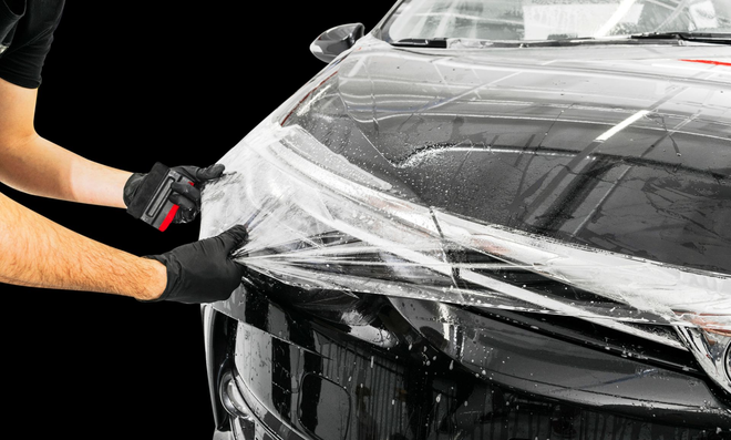 A technician in black gloves applies a clear protective film to the glossy black hood of a car.