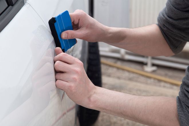 Hands use a blue squeegee with a felt edge to apply a clear protective film to a white car panel.