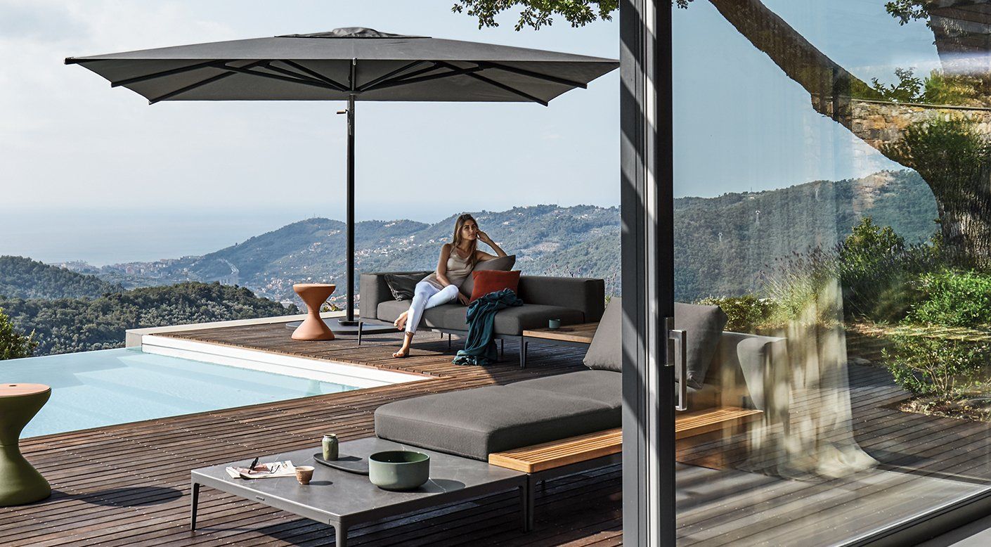 A woman is sitting on a couch under an umbrella next to a pool.