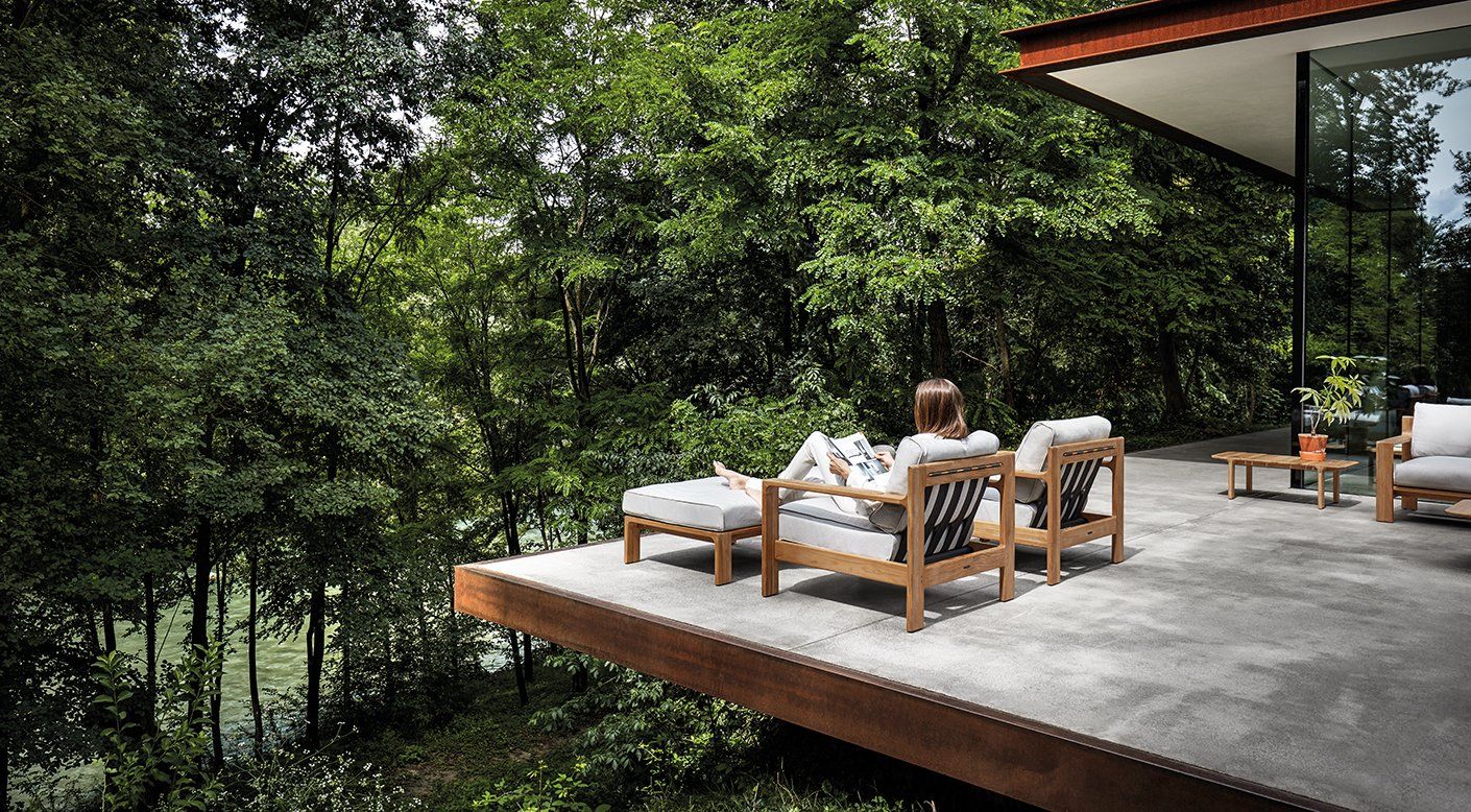 A woman is sitting on a deck in front of a house surrounded by trees.