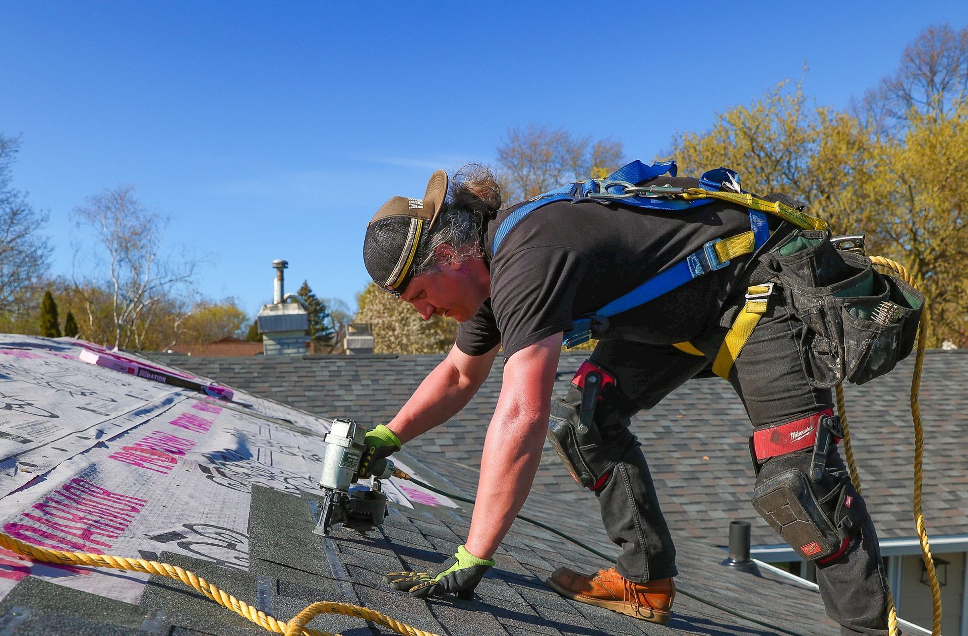 Worker using a power tool on a sloped roof, with graffiti-covered shingles under a clear blue sky