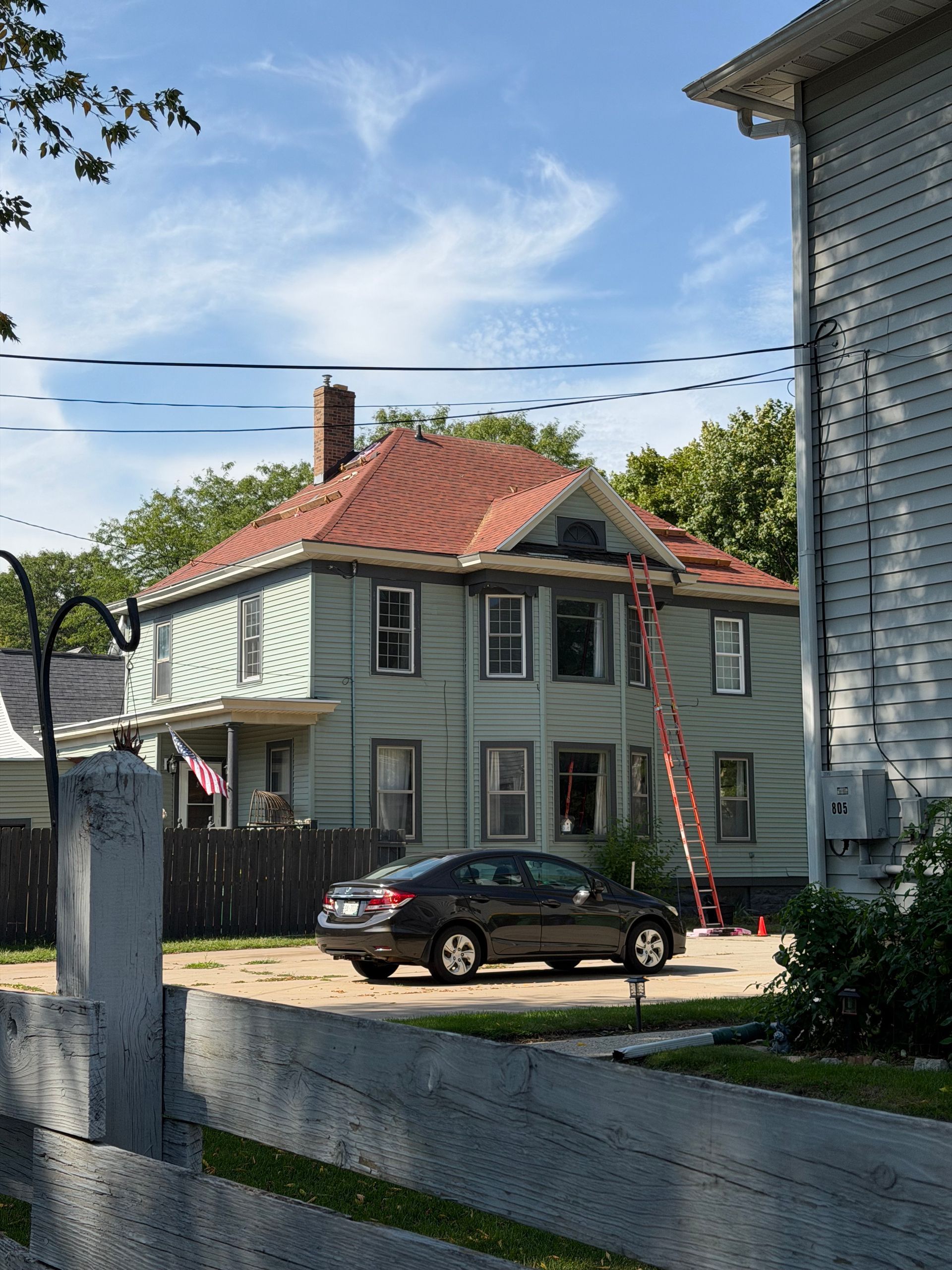 Two-story gray house with red roof, black car, and ladder in a fenced yard under a blue sky.