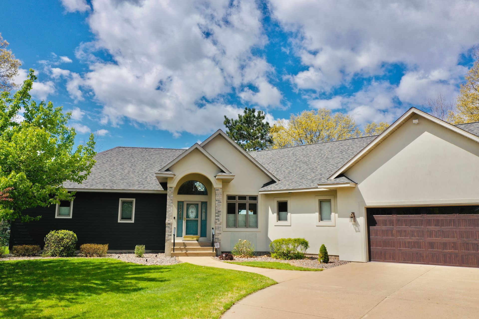 Single-story suburban house with beige siding, dark garage, and a curved driveway under a partly cloudy sky