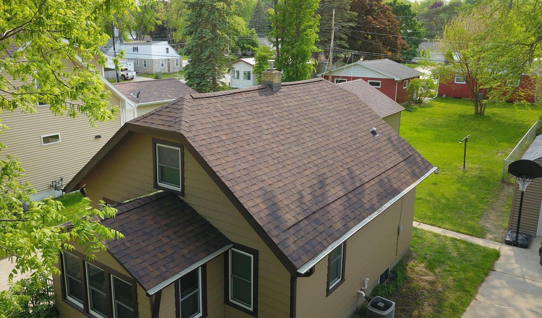 Suburban house with brown roof, green lawn, and trees in a sunny yard