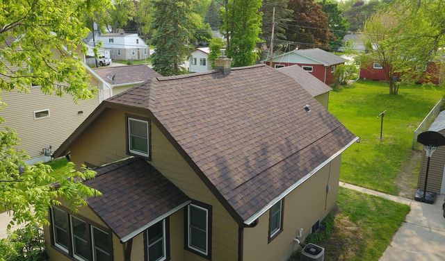 Suburban house with brown roof, green lawn, and trees in a sunny yard