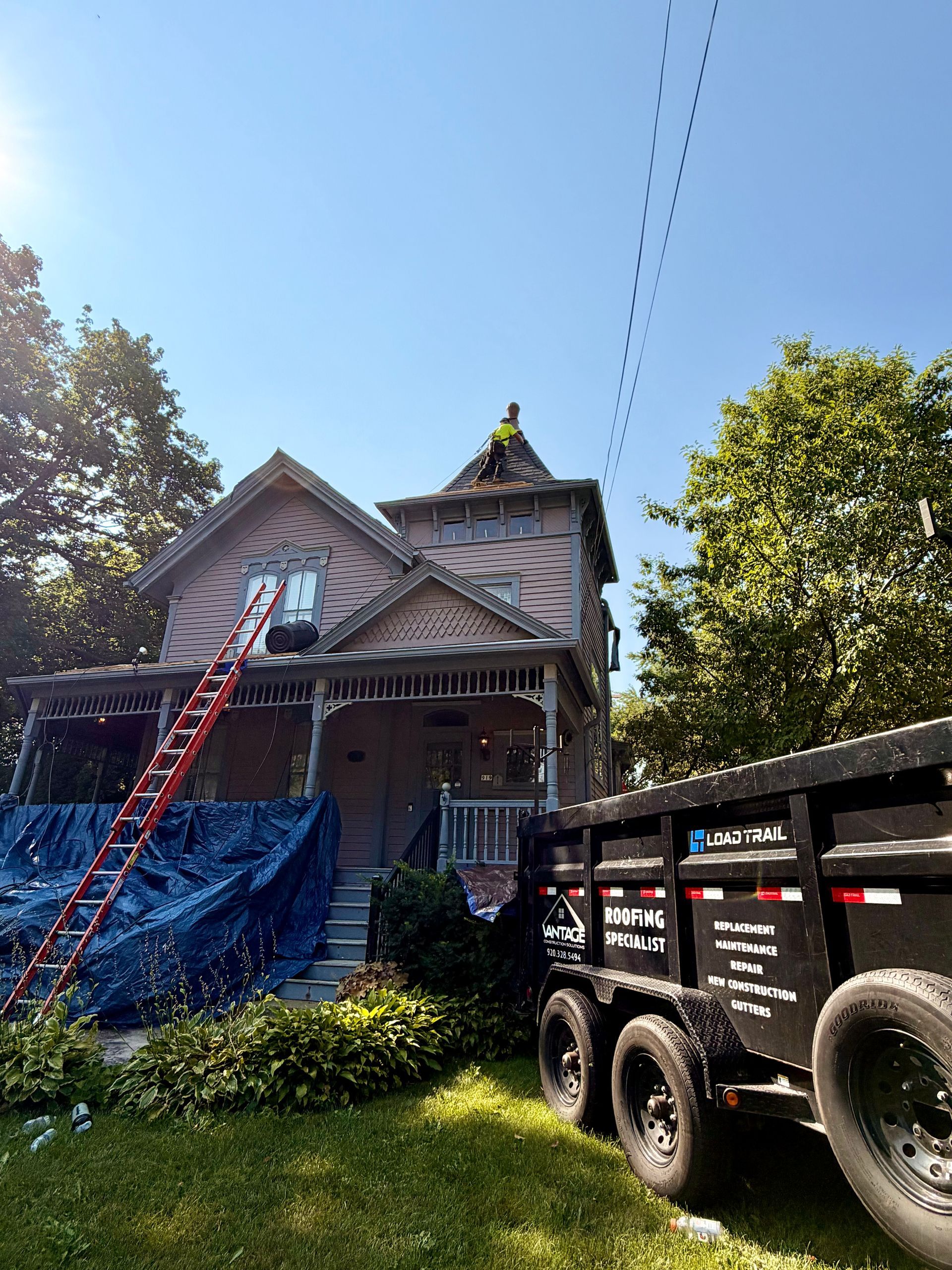 House under construction with ladders, scaffolding, and a trailer in the yard under a clear blue sky