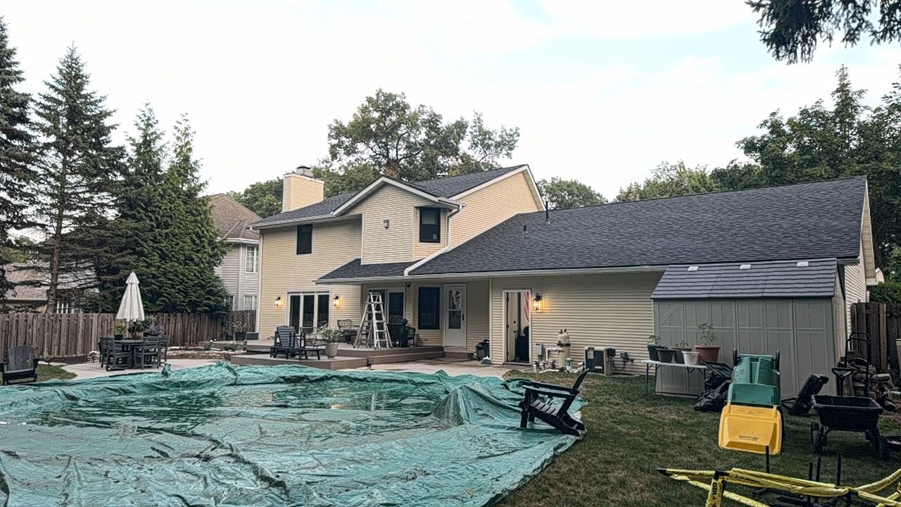 Backyard with a two-story house, covered patio, pool covered by blue tarp, and tall trees at dusk
