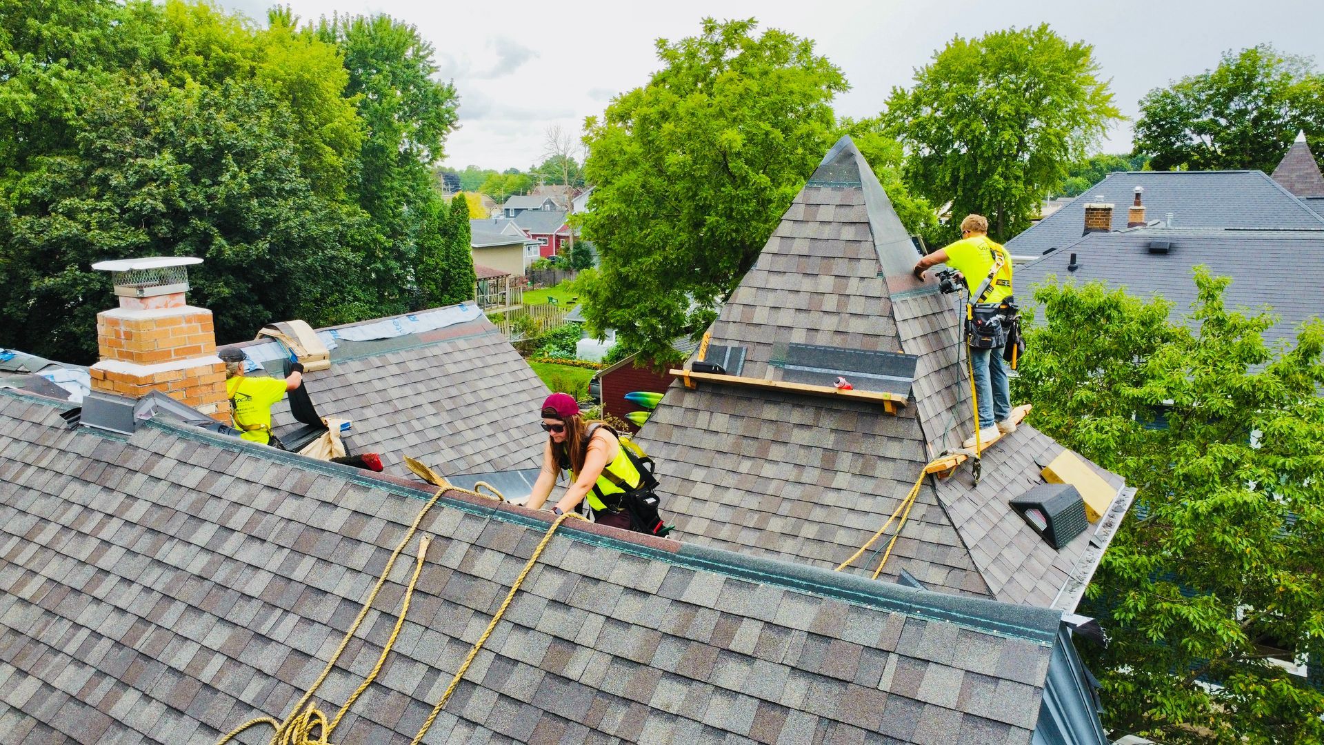 Roofers repairing a gray shingled roof beside tall green trees and neighboring houses