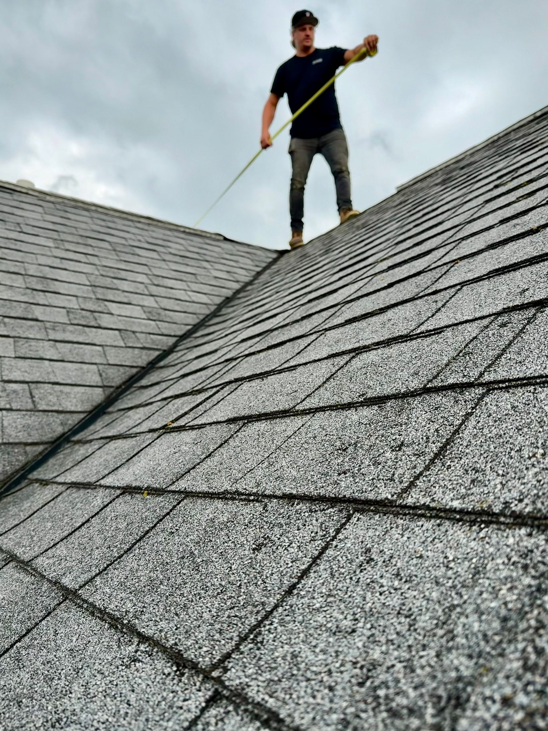 Worker on a steep shingled roof using a tool under a cloudy sky