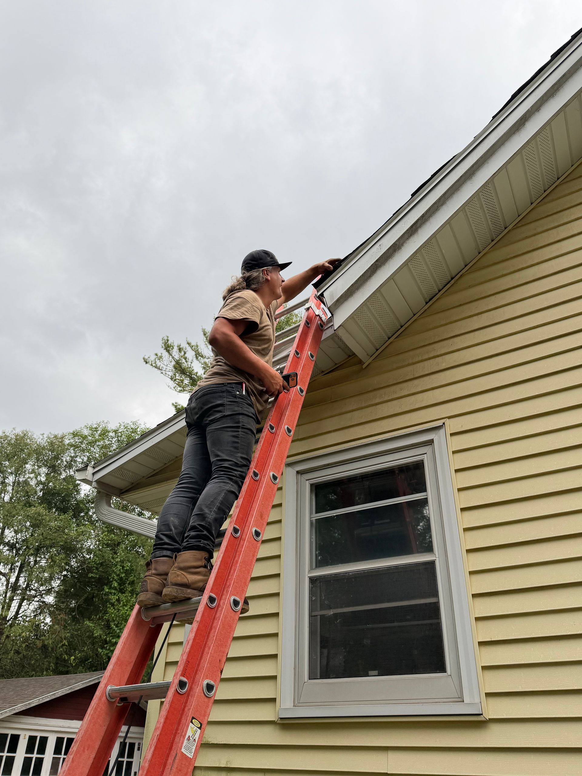 Person on a red ladder repairing a roof gutter beside a beige house