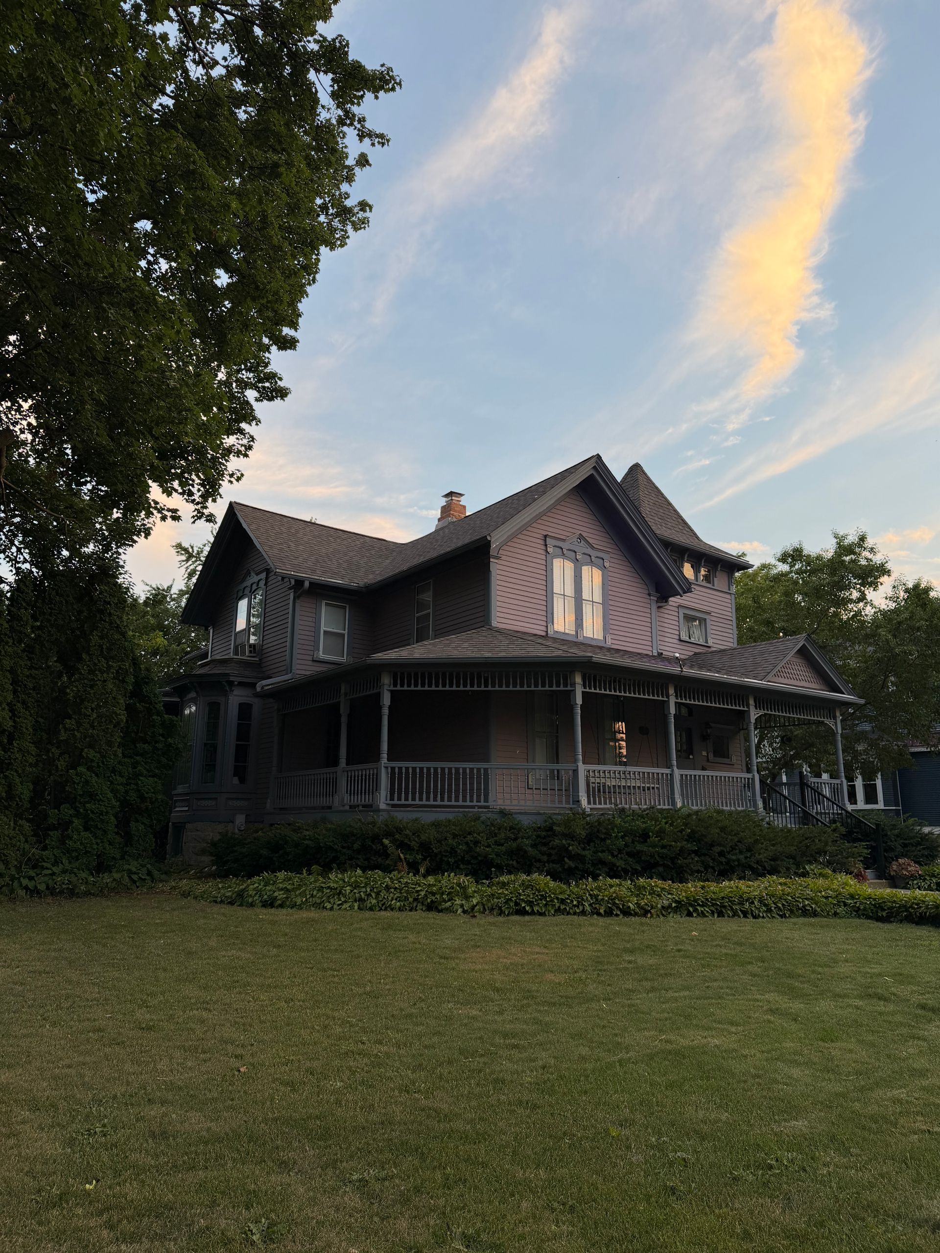 Victorian-style house with a wraparound porch, framed by trees and a grassy lawn at sunset.
