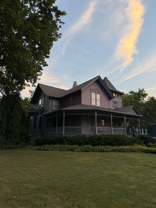 Victorian-style house with a wraparound porch, framed by trees and a grassy lawn at sunset.