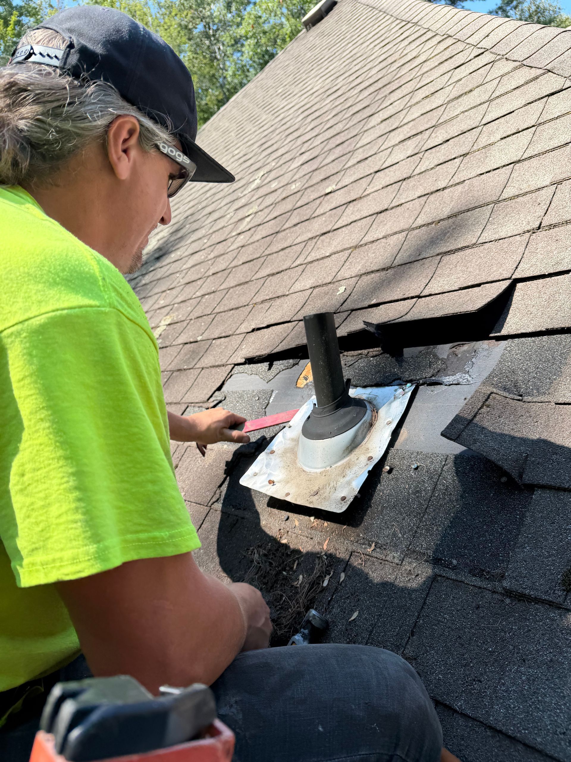 Worker inspecting a roof vent on a shingled roof, using a hand tool.